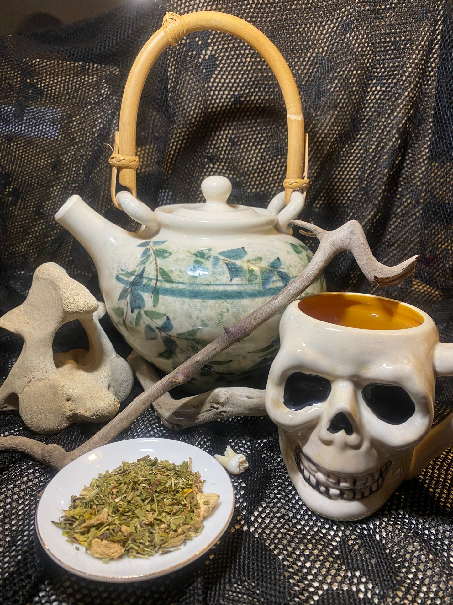 Teapot with decorative elements, skull-shaped mug, and plate of herbs on a textured surface