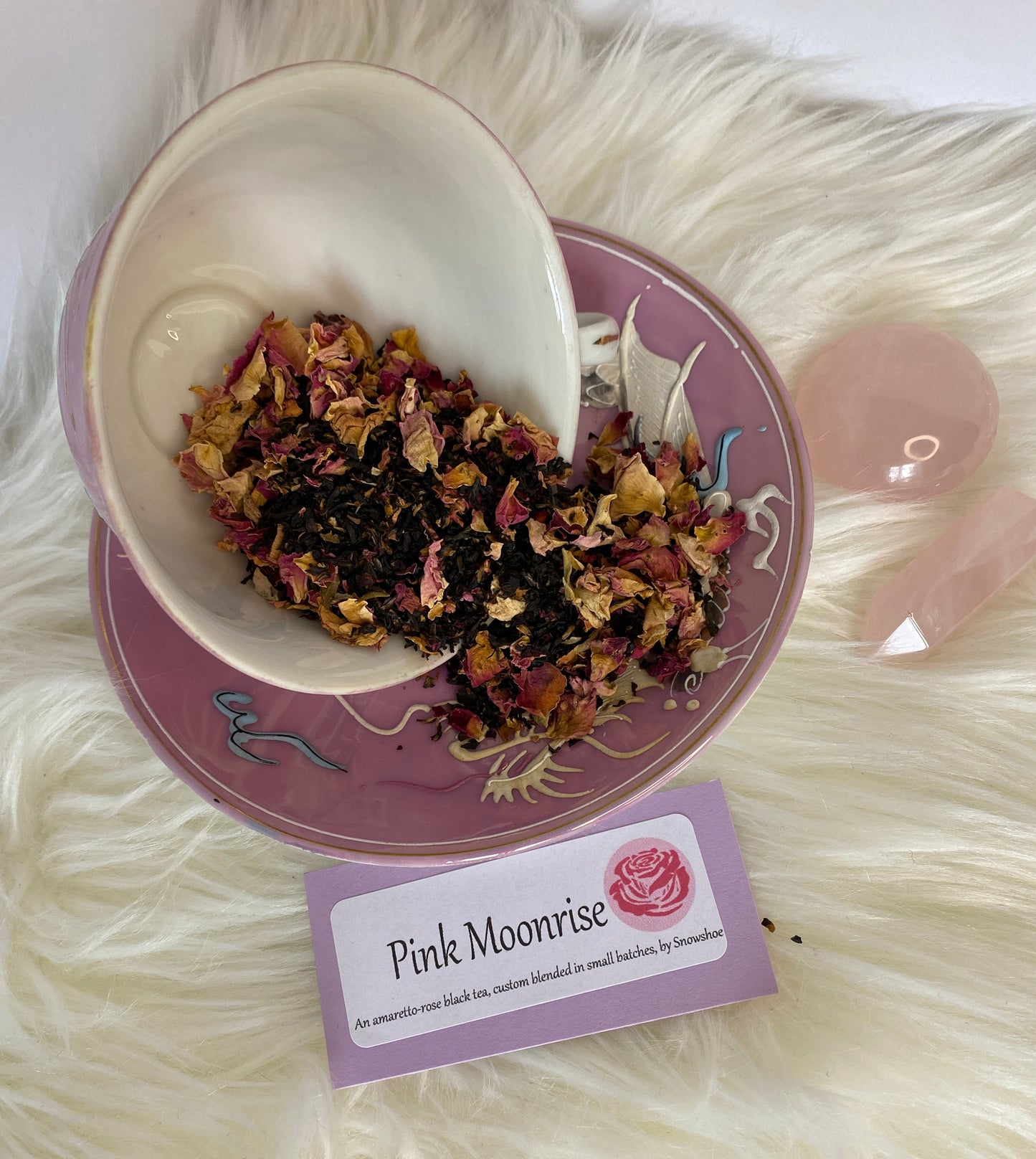 Tipped-over teacup of tea and rose petals on a decorative saucer with a 'Pink Moonrise' label on a fluffy white surface.